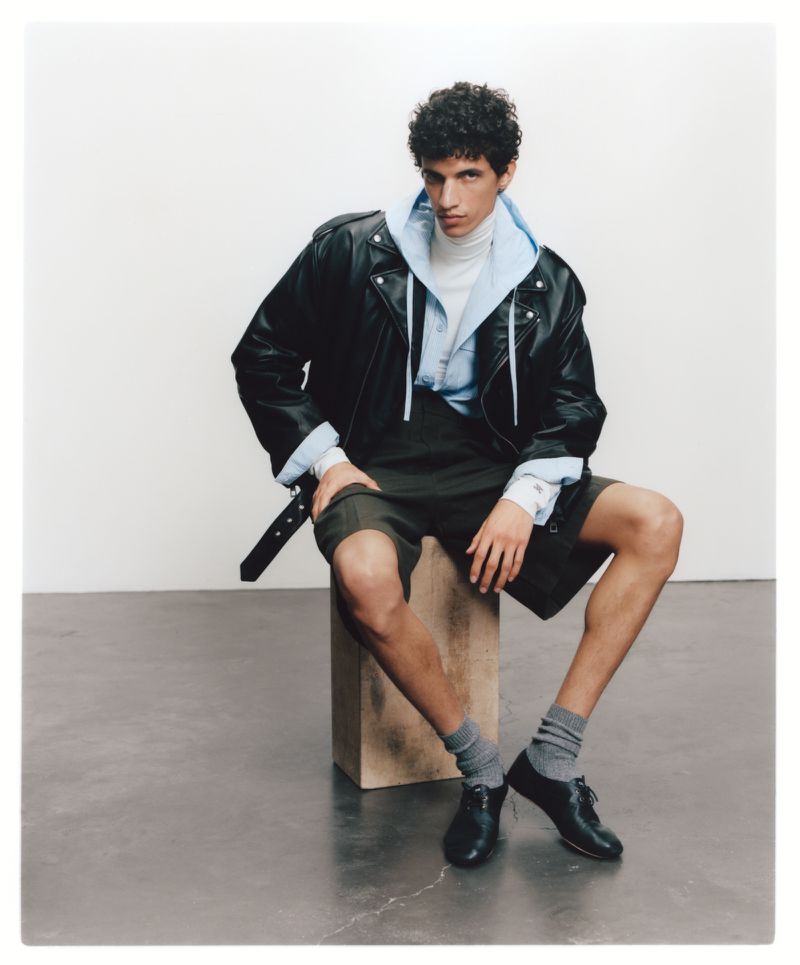 Model wearing a black leather jacket, blue hoodie, and dark shorts while seated on a wooden block in a studio.