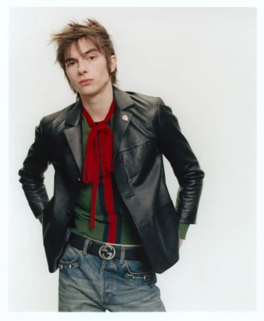 Model with messy hair wearing a black leather jacket, green striped top, and red necktie against a white backdrop.