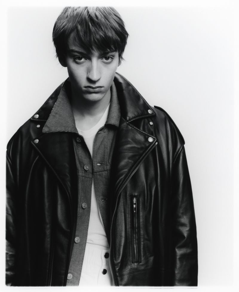 Monochrome portrait of a young male model wearing an oversized black leather jacket against a white background