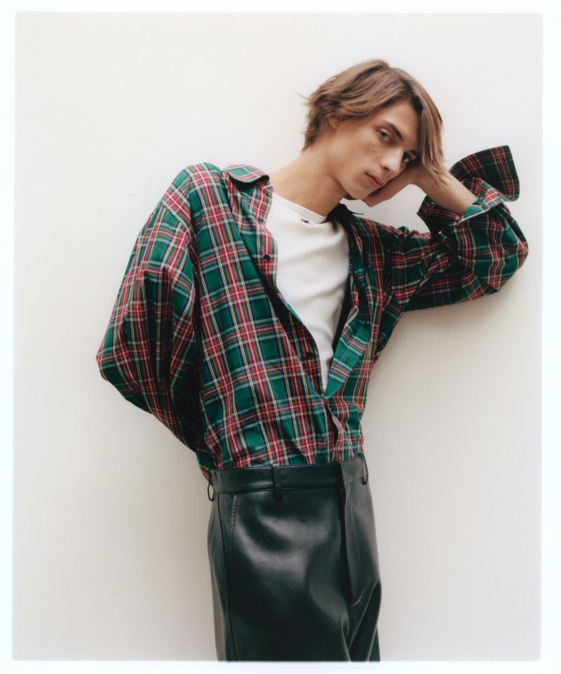 Model wearing a plaid shirt and leather trousers posing against a white seamless background for Behind the Blinds.