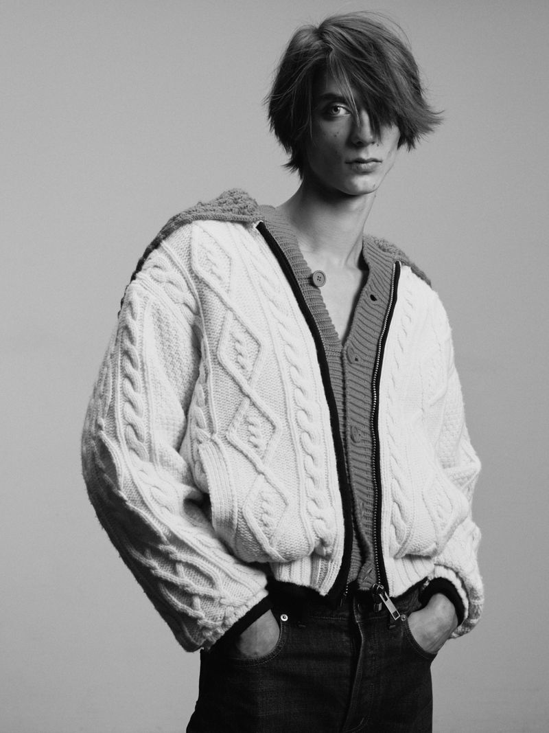 Young model with dark hair wearing a white cable-knit cardigan against a plain light grey studio background.