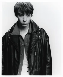 Monochrome portrait of a young male model wearing an oversized black leather jacket against a white background