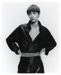 Model wearing a black leather shirt and trousers with hands on hips against a white studio background