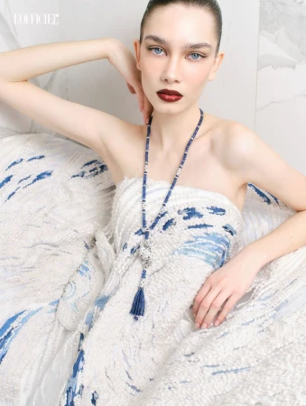 Close-up portrait of a woman wearing a strapless textured white and blue dress and a long beaded necklace for L'Officiel Belgium.