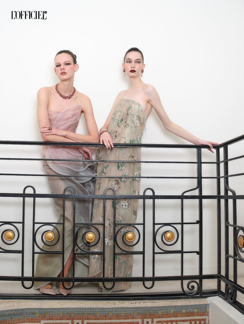 Two female models in strapless evening gowns posing behind a decorative black metal railing for L'Officiel Belgium.
