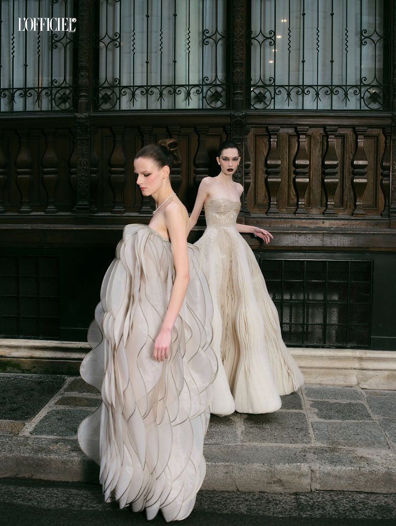 Two female models in sculptural gowns posing outdoors in front of a dark wooden architectural facade.