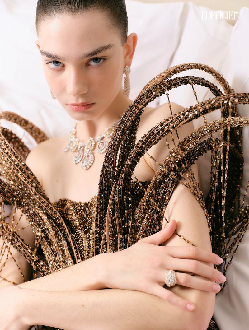 Close-up of a young woman in an embellished brown dress and jewelry lying on a white surface, portrait style, fashion editorial.