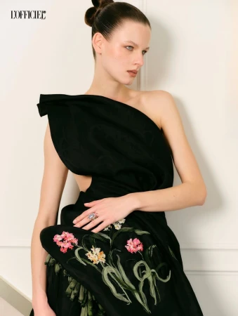 Model in a black asymmetric dress with floral embroidery posing against a white studio wall