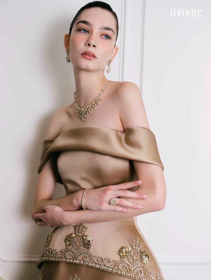 Model in off-the-shoulder gold satin gown with intricate beadwork wearing a matching necklace and earrings against a white background