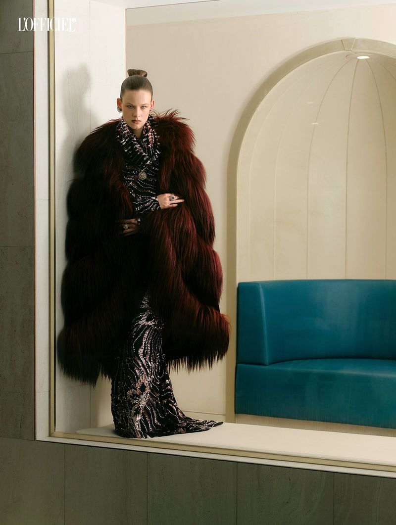 Model wearing a voluminous burgundy fur coat and dark patterned gown standing inside an architectural interior.
