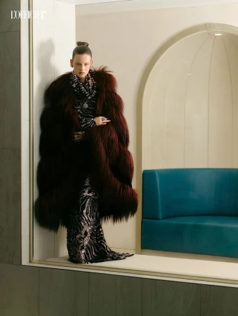 Model wearing a voluminous burgundy fur coat and dark patterned gown standing inside an architectural interior.
