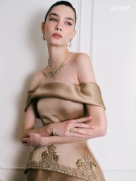 Model in off-the-shoulder gold satin gown with intricate beadwork wearing a matching necklace and earrings against a white background