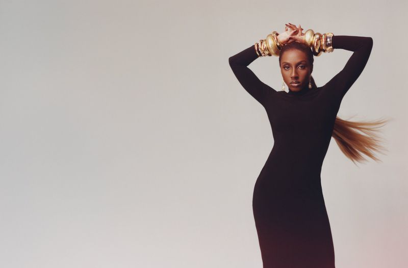 Ziwe Fumudoh in a black dress wearing thick gold bangles against a neutral background for Love Magazine.