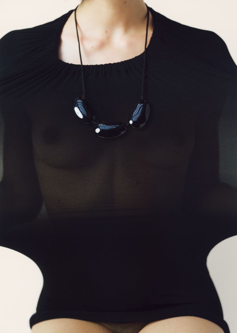 Model wearing a black sheer top and a large dark pebble necklace against a plain background