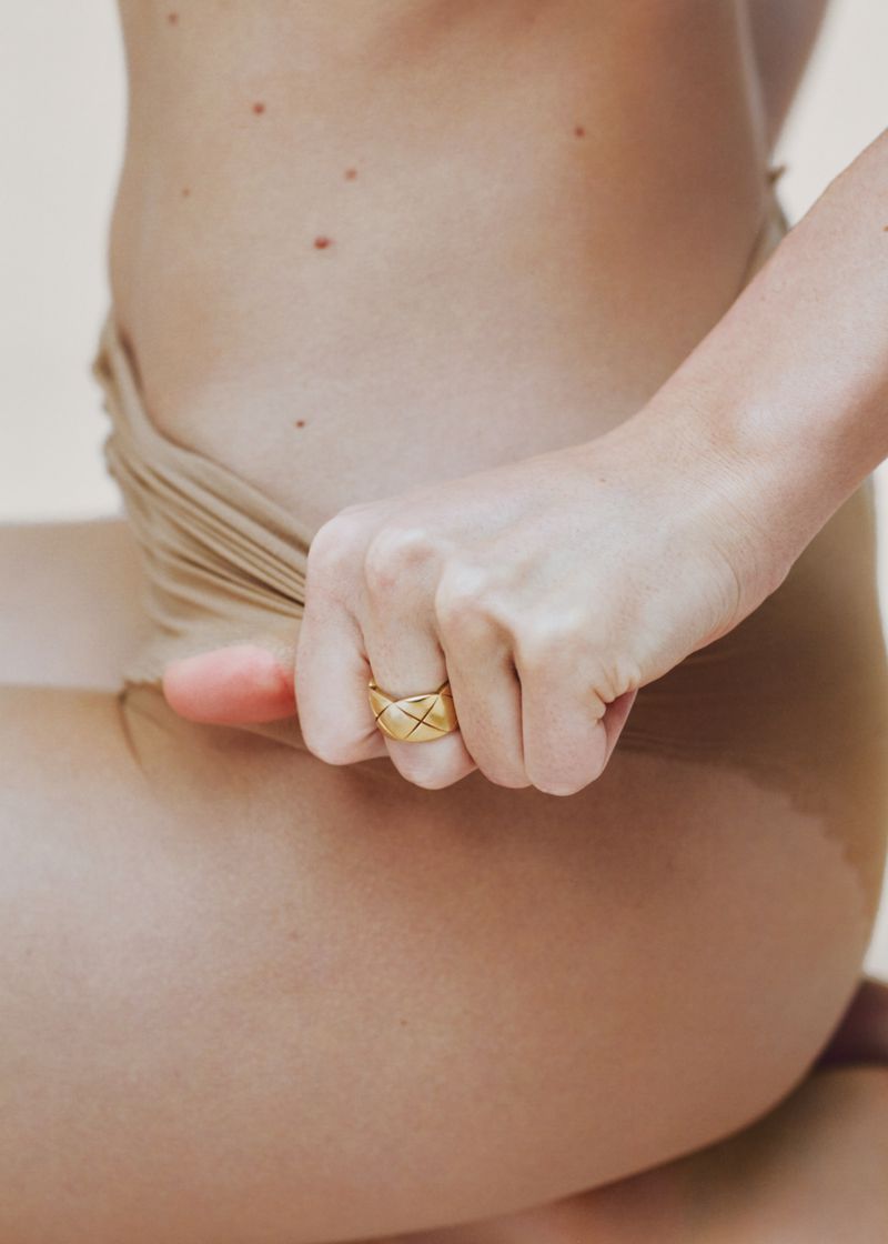Close-up of a person wearing a gold quilted ring and beige swimwear against a neutral background.