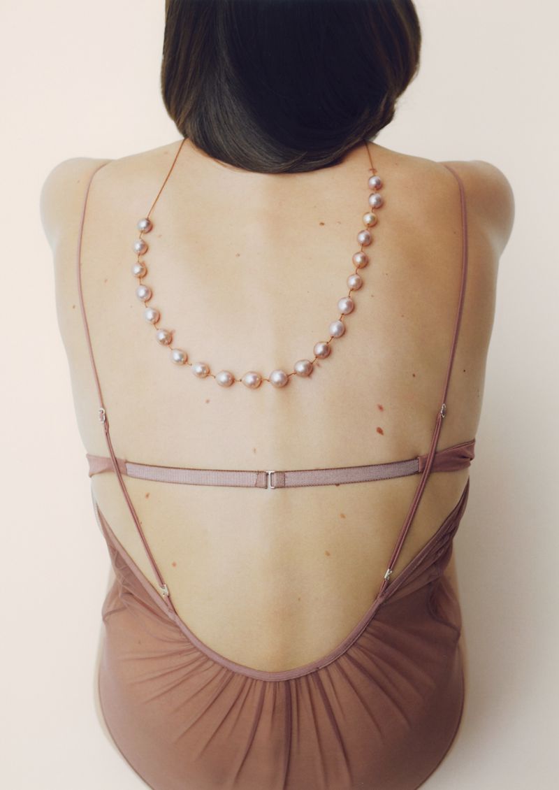 Rear view of a woman wearing a low-backed garment and a pearl necklace against a plain light background.