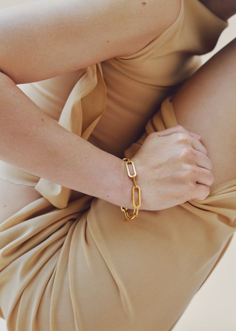 Close-up of a person wearing a gold chain link bracelet and beige clothing against a neutral background
