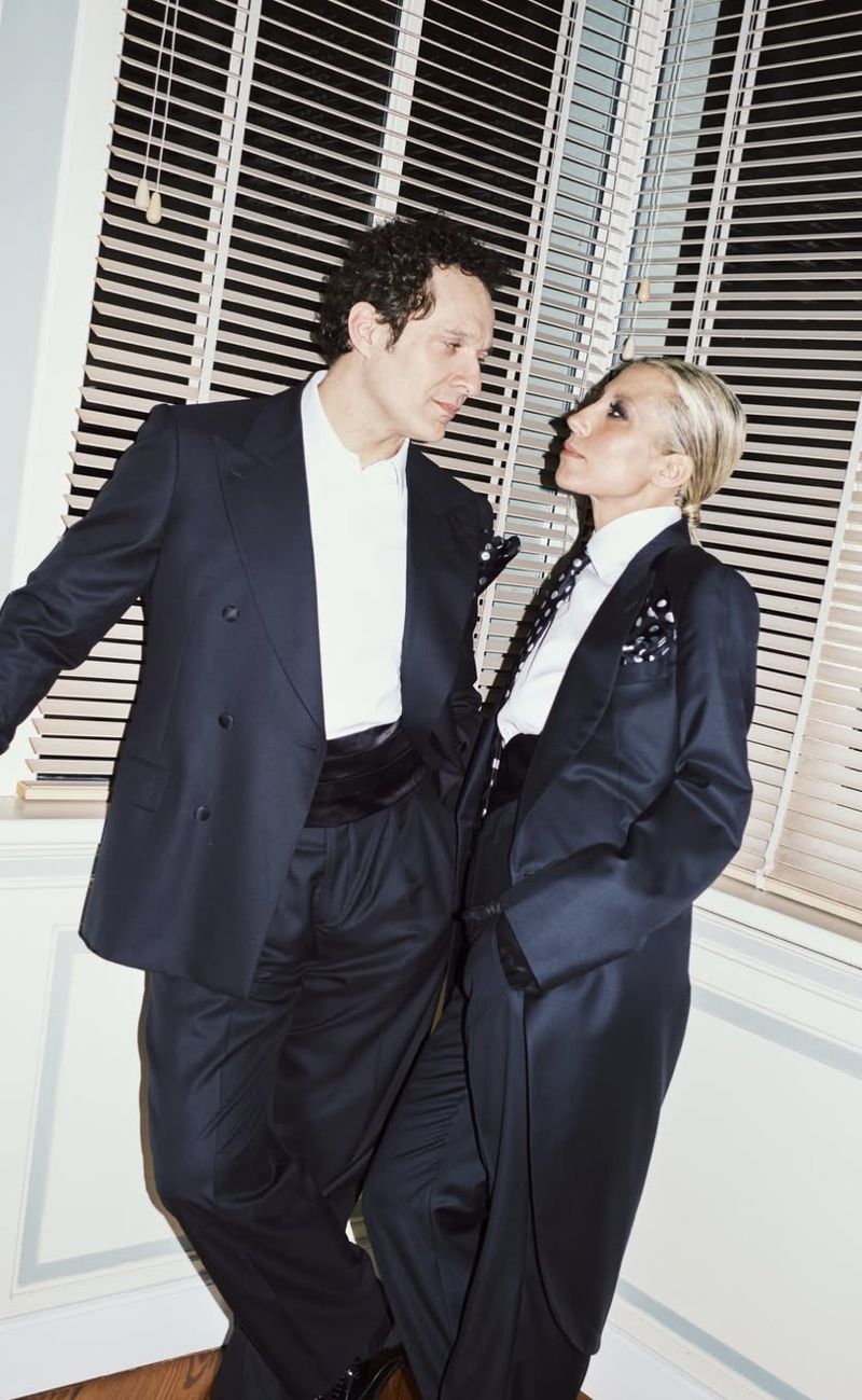 Elegant couple in formal wear poses indoors against a window with blinds. The woman wears a tuxedo; the man sports a suit and cummerbund.