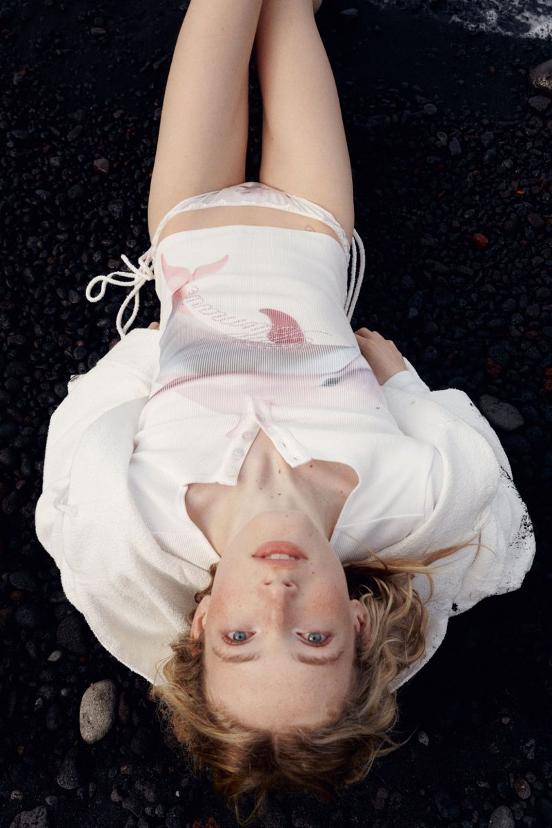 Woman in white swimsuit and shirt lies on black pebbles at the beach, an overhead shot showcasing fashion and swimwear.