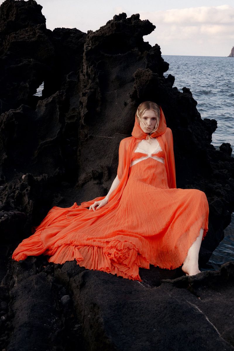 Woman in orange gown sits on black rocks near ocean. Fashion shot with model in gown, dark rocks, ocean view, neutral expression.