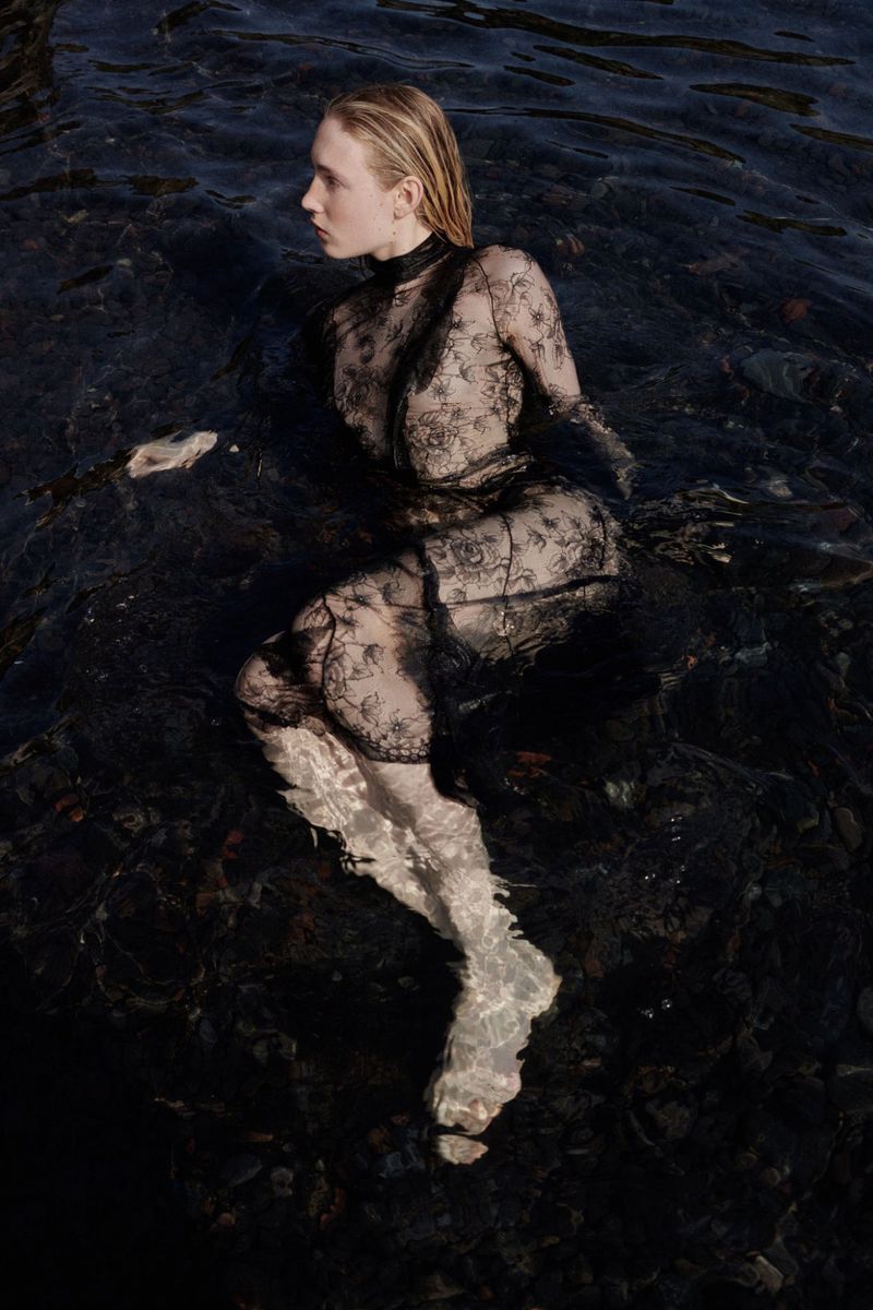 A blonde woman in a lace dress is waist-deep in dark, rippling water in a fashion-style photograph with shallow depth of field and muted lighting.