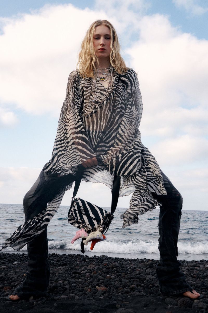 Fashion shot of a blonde adult female posing on a black sand beach wearing a zebra-print tunic, pants, and a matching zebra bag.