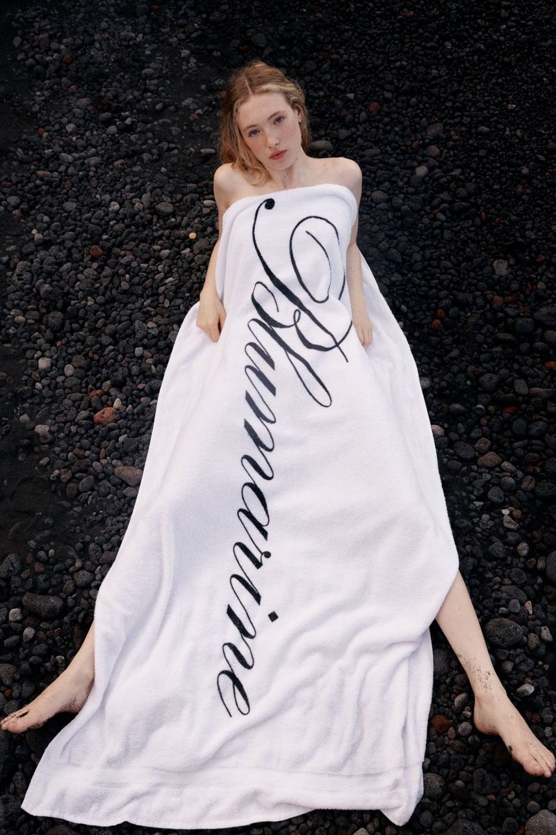 Young woman poses wrapped in a white logo towel on a black pebble beach, eye-level shot, natural lighting, fashion editorial photography.