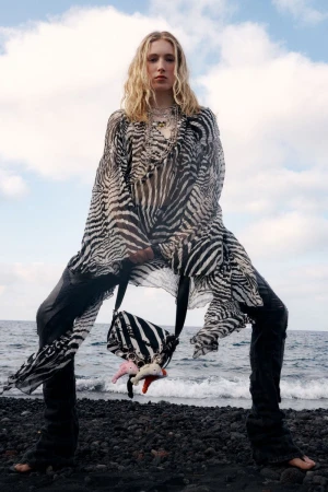 Model posing in a sheer zebra-print draped top and black pants on a dark rocky beach holding a matching printed crossbody bag.