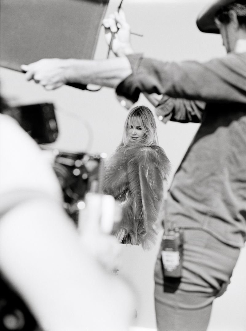 Black and white behind-the-scenes fashion shoot showing crew members and a blonde female model in a textured coat.