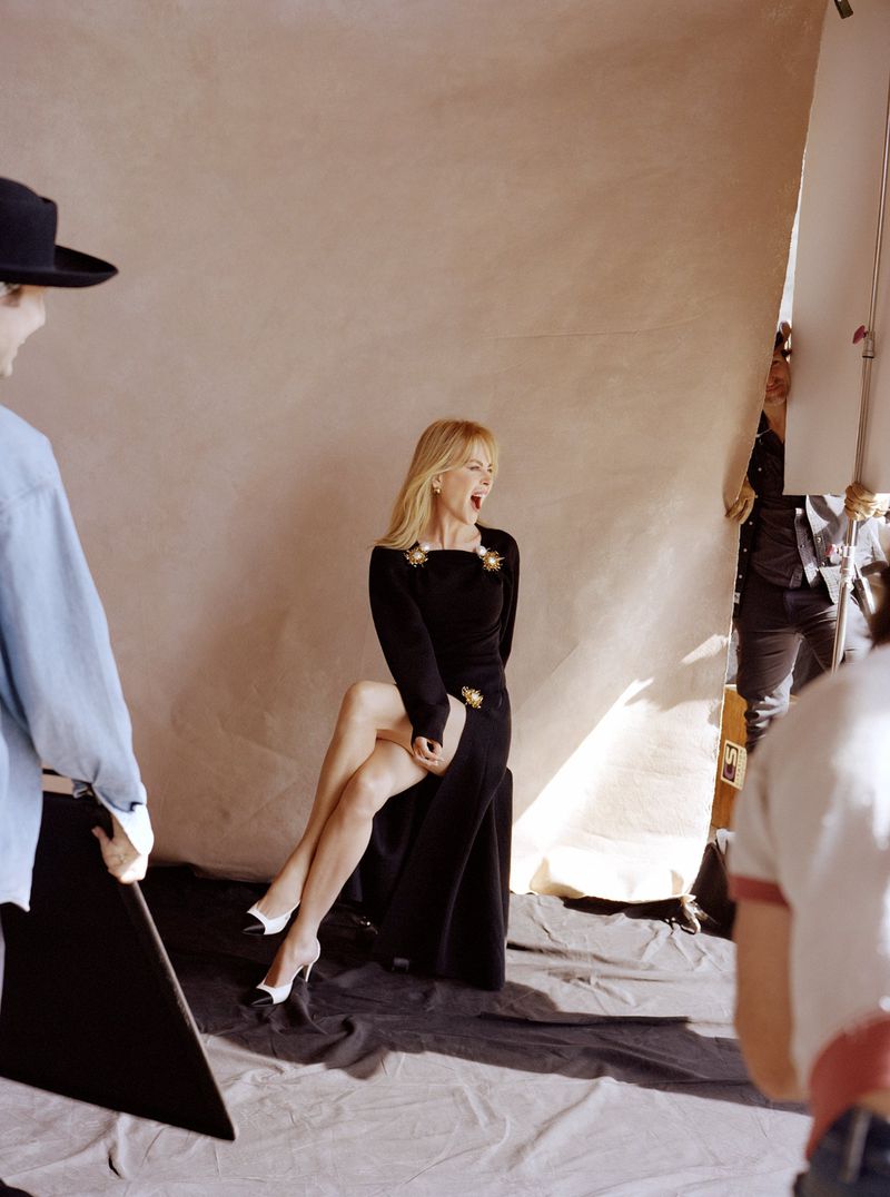 Nicole Kidman seated in a black dress laughing during a photoshoot with crew members visible around her