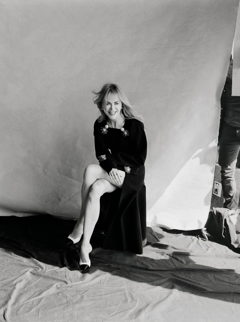 Nicole Kidman sitting on a stool against a white seamless backdrop wearing a black dress and white heels in a behind the scenes portrait