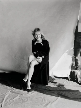 Nicole Kidman sitting on a stool against a white seamless backdrop wearing a black dress and white heels in a behind the scenes portrait