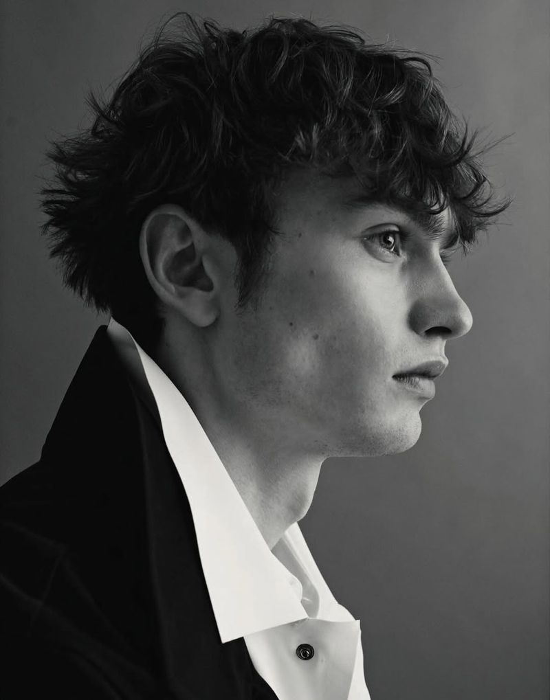 Profile portrait of a young man with dark curly hair wearing a white collared shirt and dark jacket against a grey background.