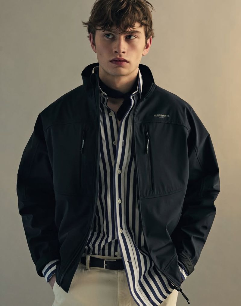 Model Kian Cleator wearing a dark jacket over a striped shirt and pale trousers against a plain studio backdrop.