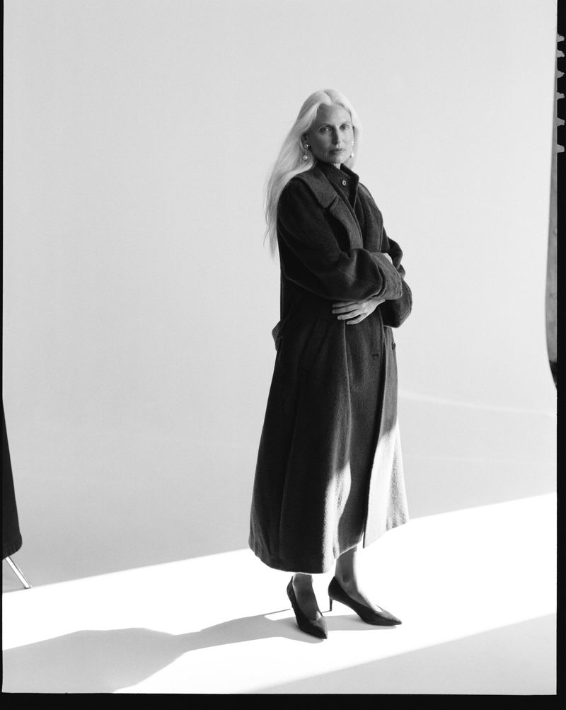 Model wearing a dark long coat and heels with arms crossed against a white background for P. Johnson Femme Fall 26