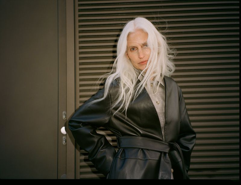 Model with long white hair wearing a black belted coat against a slatted metallic background