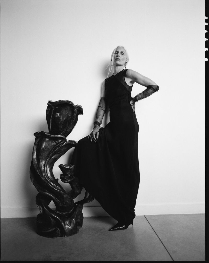 Model in long black sleeveless dress posing next to a sculptural dark art piece against a plain studio wall