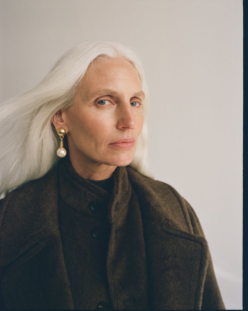 Model with long white hair wearing a dark brown coat and pearl drop earrings against a neutral grey wall