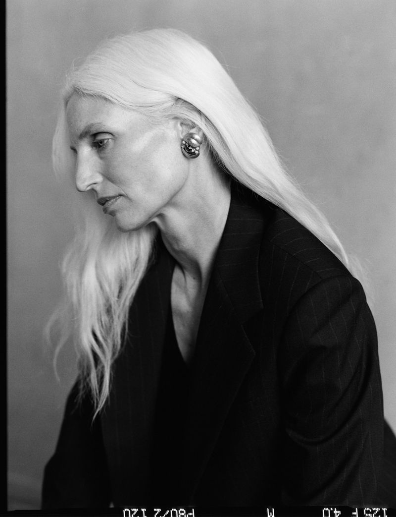 Monochrome portrait profile of a woman with long white hair wearing a dark blazer and statement earrings