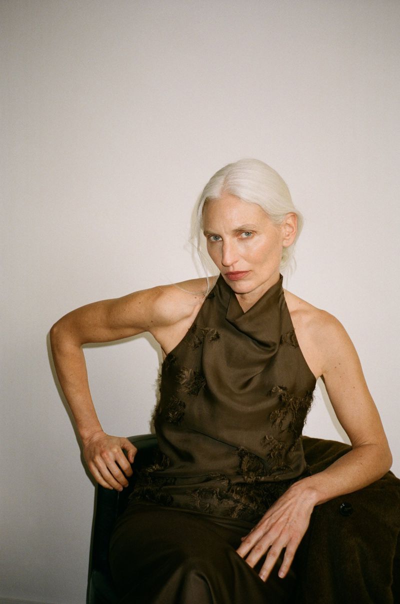 Model with white hair wearing a dark halter top sitting against a plain white wall