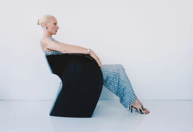 Model Maggie Maurer seated in a black modern chair wearing a blue textured gown against a white background