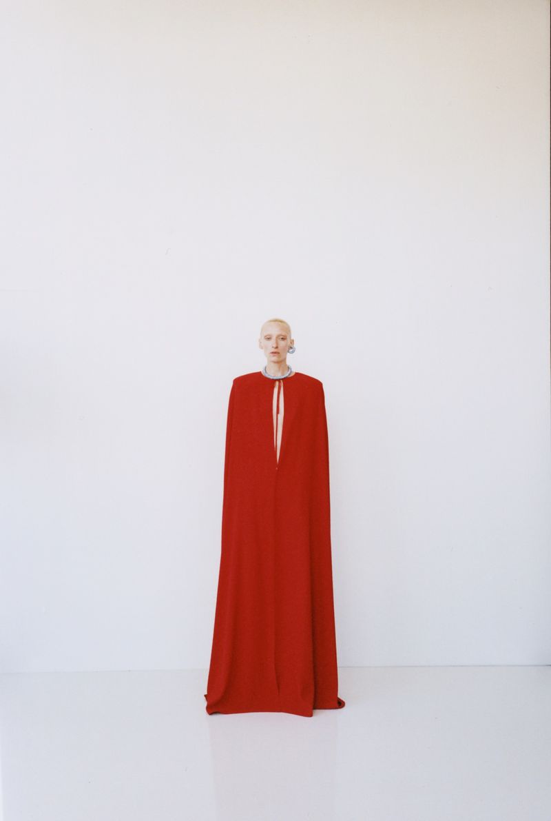 Model wearing a long red cape gown standing against a plain white background for The New Arrivals lookbook project
