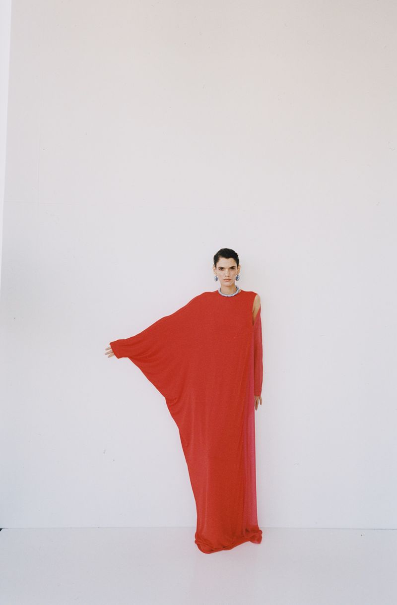 Model wearing a flowing red dress standing against a plain white studio backdrop for The New Arrivals project.