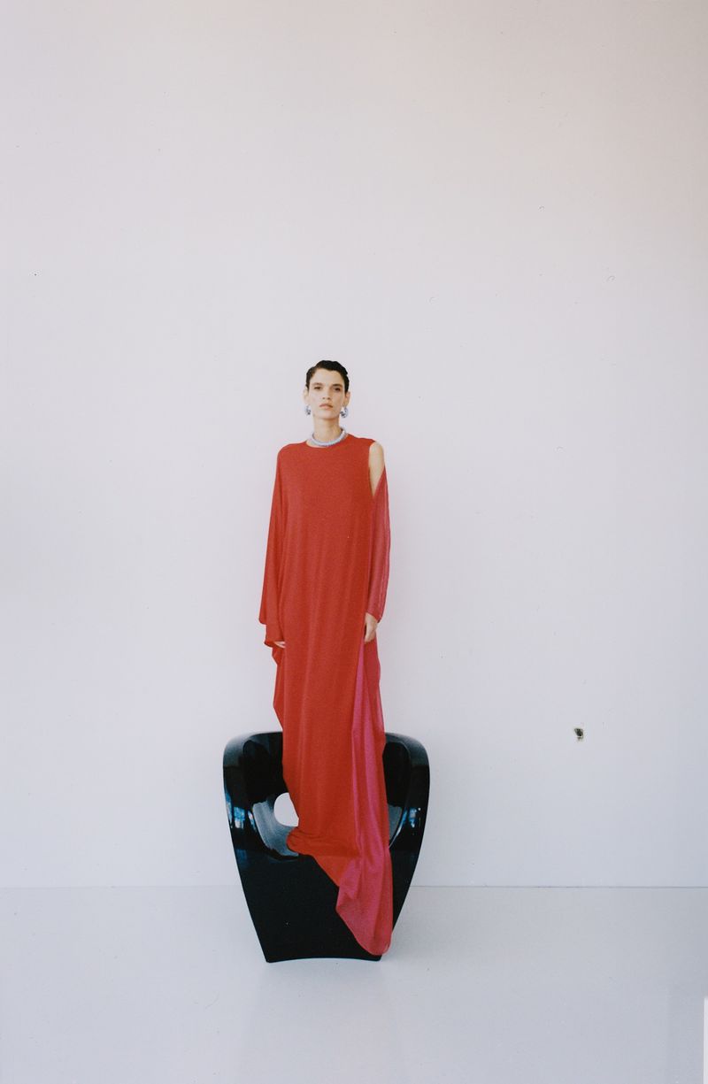 Model in a long red dress standing on a black chair against a white wall background