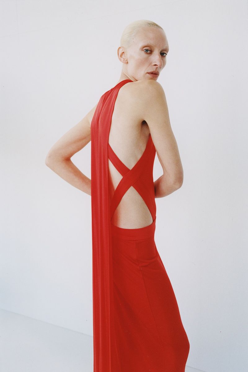 Model with bleached hair wearing a backless red dress against a plain white studio background