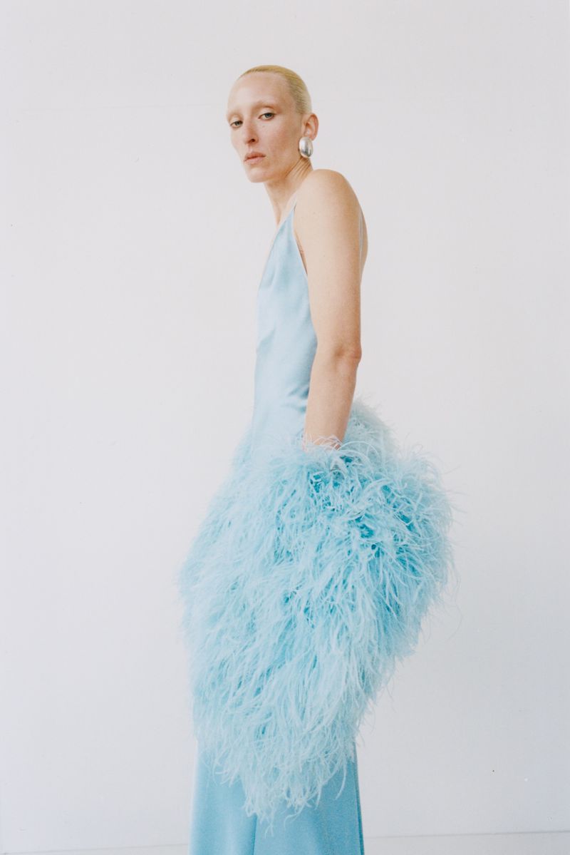 Model wearing a light blue slip dress adorned with fluffy feathers against a plain white studio wall.