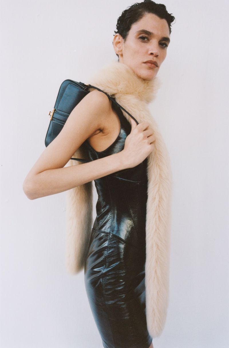 Model with short dark hair wearing a glossy black dress and furry cream scarf holding a black shoulder bag against white