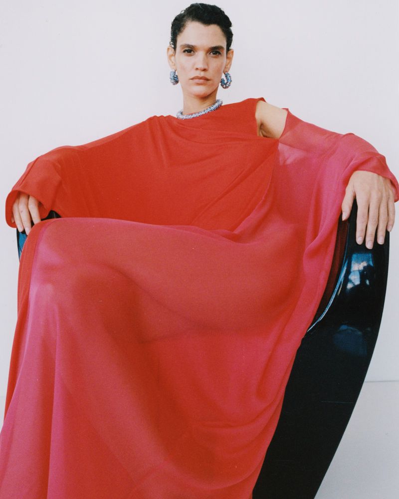 Model seated in a black chair wearing a vibrant red flowing dress and statement hoop earrings against a white background