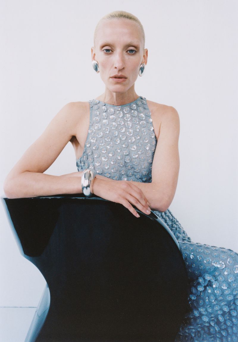 Model wearing a textured blue sleeveless dress and silver jewelry seated behind a black curved chair against a white background