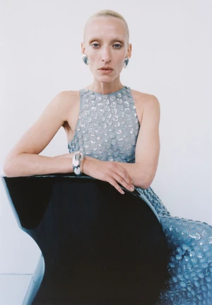 Model wearing a textured blue sleeveless dress and silver jewelry seated behind a black curved chair against a white background